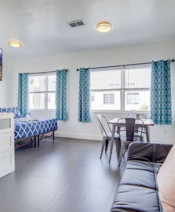 The image shows a modern room with a bed, sofa, small table with chairs, TV, and wall art, featuring blue curtains and natural light.