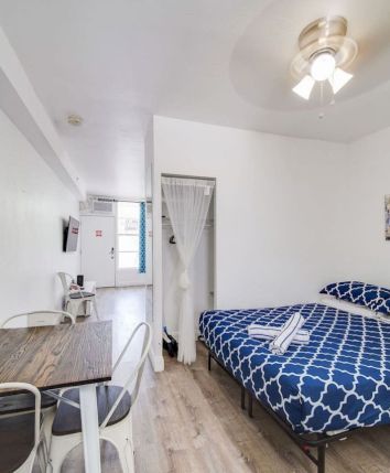 A modern bedroom with a blue-patterned bed, table with chairs, pineapple decor, wall art, and a ceiling fan, in a bright and minimal design.