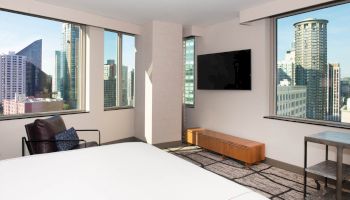 A modern hotel room with large windows, a bed, flat-screen TV, city views, and contemporary furniture, creating a bright, urban ambiance.
