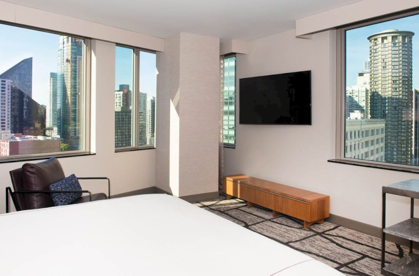 A modern hotel room with large windows, a bed, flat-screen TV, city views, and contemporary furniture, creating a bright, urban ambiance.