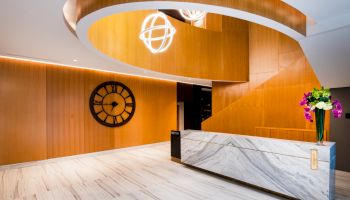 A modern lobby with wooden walls, a large clock, marble reception desk, spiral design, and flowers on the counter, under bright lighting.