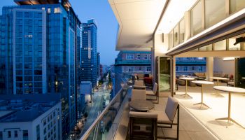 A modern rooftop terrace with seating overlooks a city street lined with tall buildings, illuminated under a twilight sky.