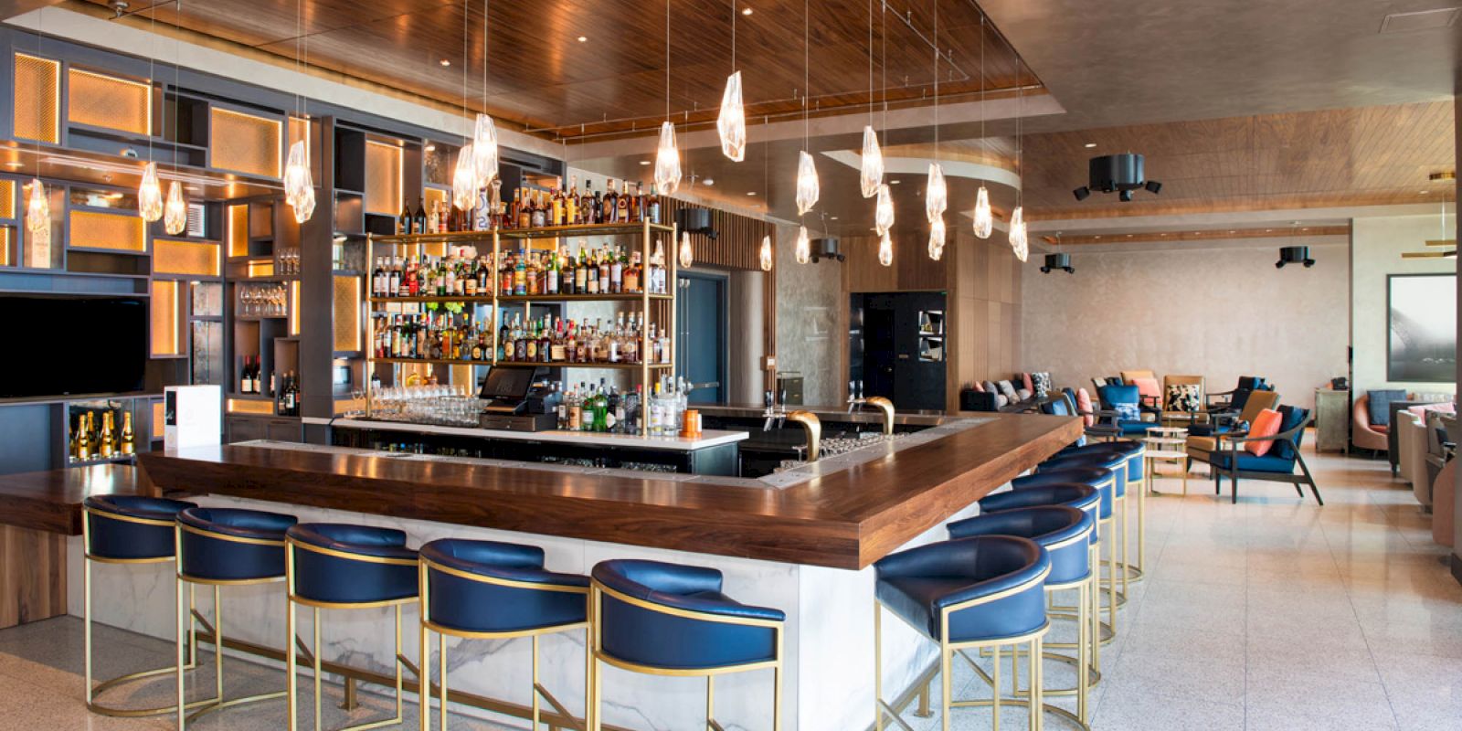 The image shows a stylish bar with blue stools, pendant lights, and a variety of bottles on shelves. The decor is modern and inviting.