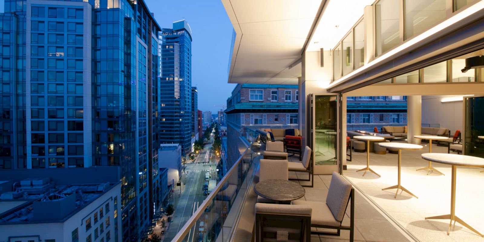 The image shows a modern balcony with seating overlooking a cityscape at dusk, featuring high-rise buildings and a lit street below.