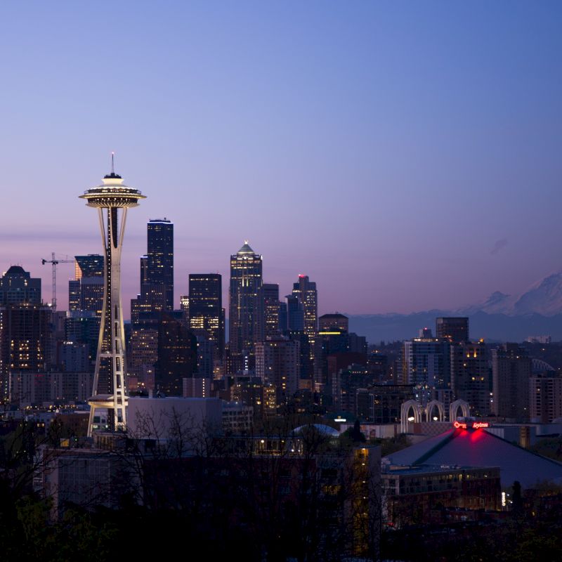 A cityscape at dusk with a prominent tower and mountains in the background, likely depicting Seattle with the Space Needle in view.