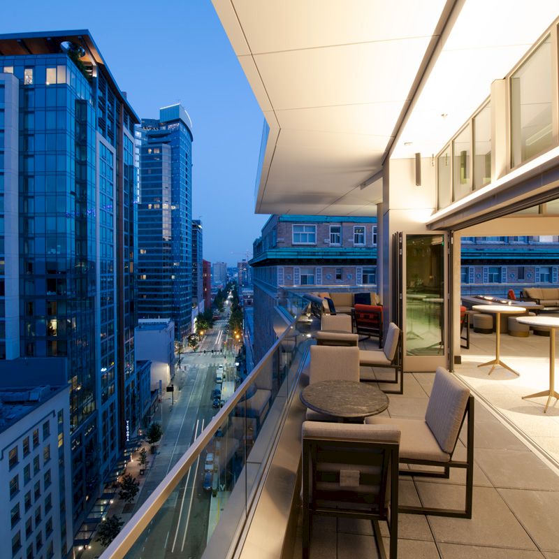 Rooftop view of a modern cityscape at dusk, featuring high-rise buildings, stylish outdoor seating, and well-lit interiors.