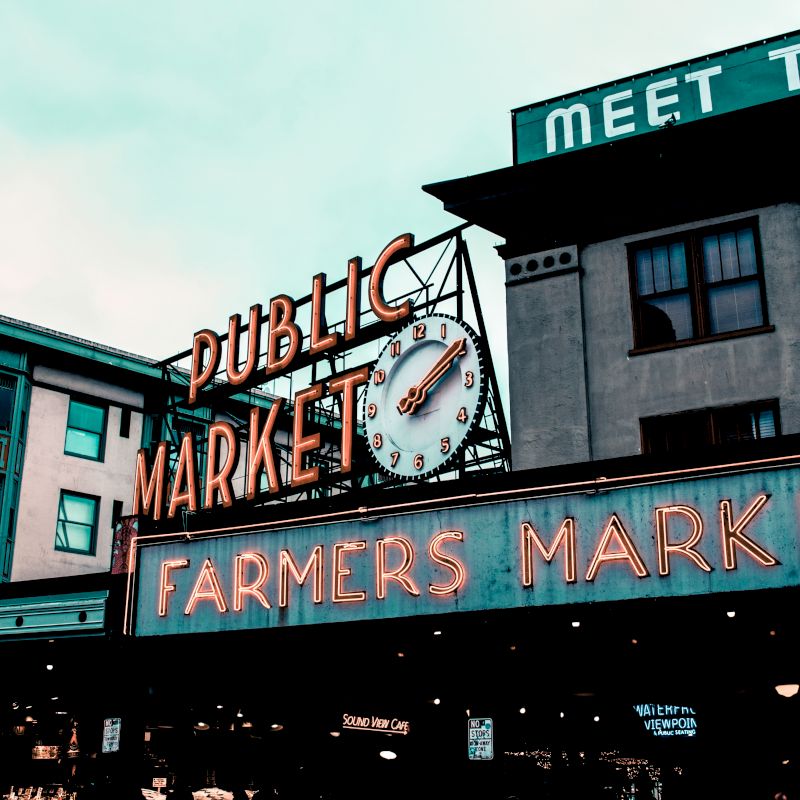 The image shows an iconic neon sign for a public market and farmers market, with buildings in the background.