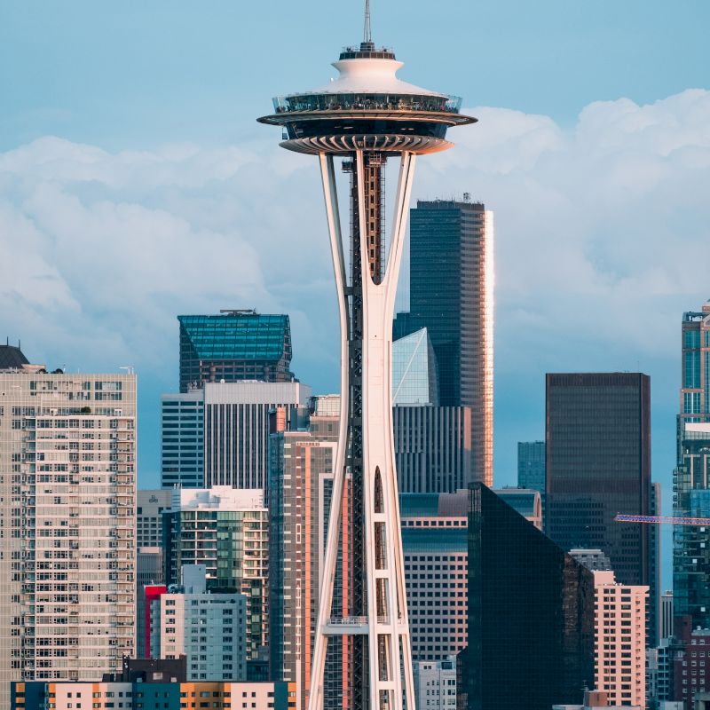 The image shows the Space Needle in Seattle, surrounded by city skyscrapers against a cloudy blue sky backdrop.