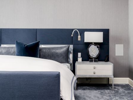 A modern bedroom with a blue and white theme features a bed, pillows, a nightstand with a lamp, and a decorative mirror above.
