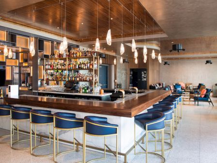 The image shows a modern bar with a central counter, blue stools, a variety of bottles, and stylish lighting fixtures.