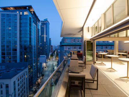 The image shows a modern rooftop terrace with seating, overlooking a city street and buildings during twilight.
