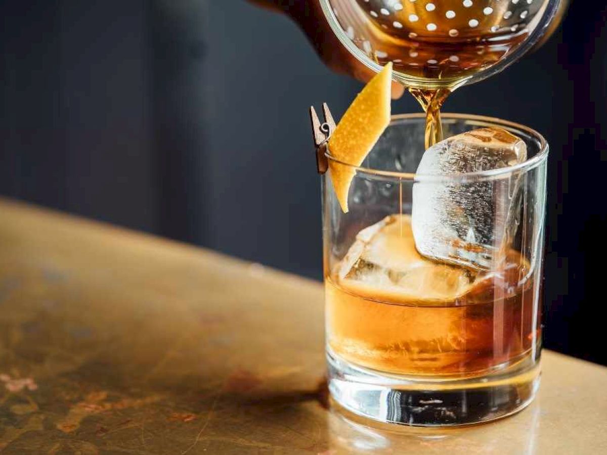 A bartender pours a cocktail into a glass with ice and a citrus twist garnish on a counter.