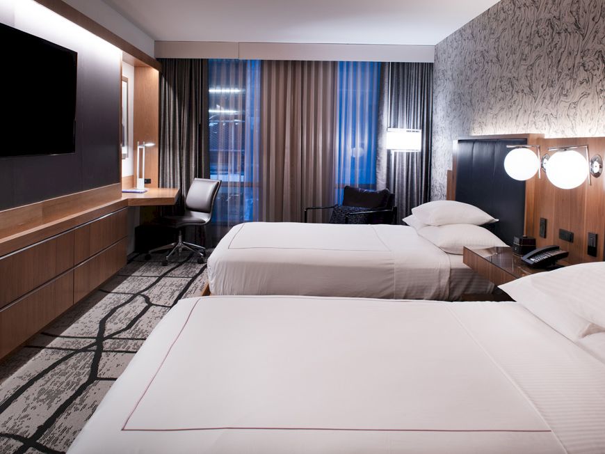 A modern hotel room with two beds, a large TV, desk, and chair. The decor features neutral tones and ambient lighting.