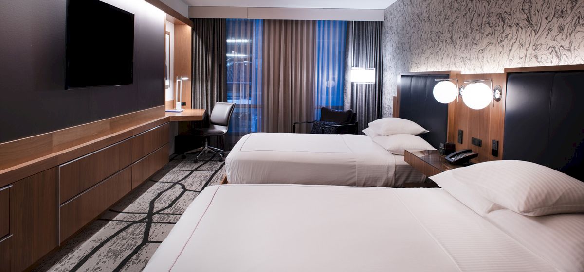 A modern hotel room with two beds, a large TV, desk, and chair. The decor features neutral tones and ambient lighting.