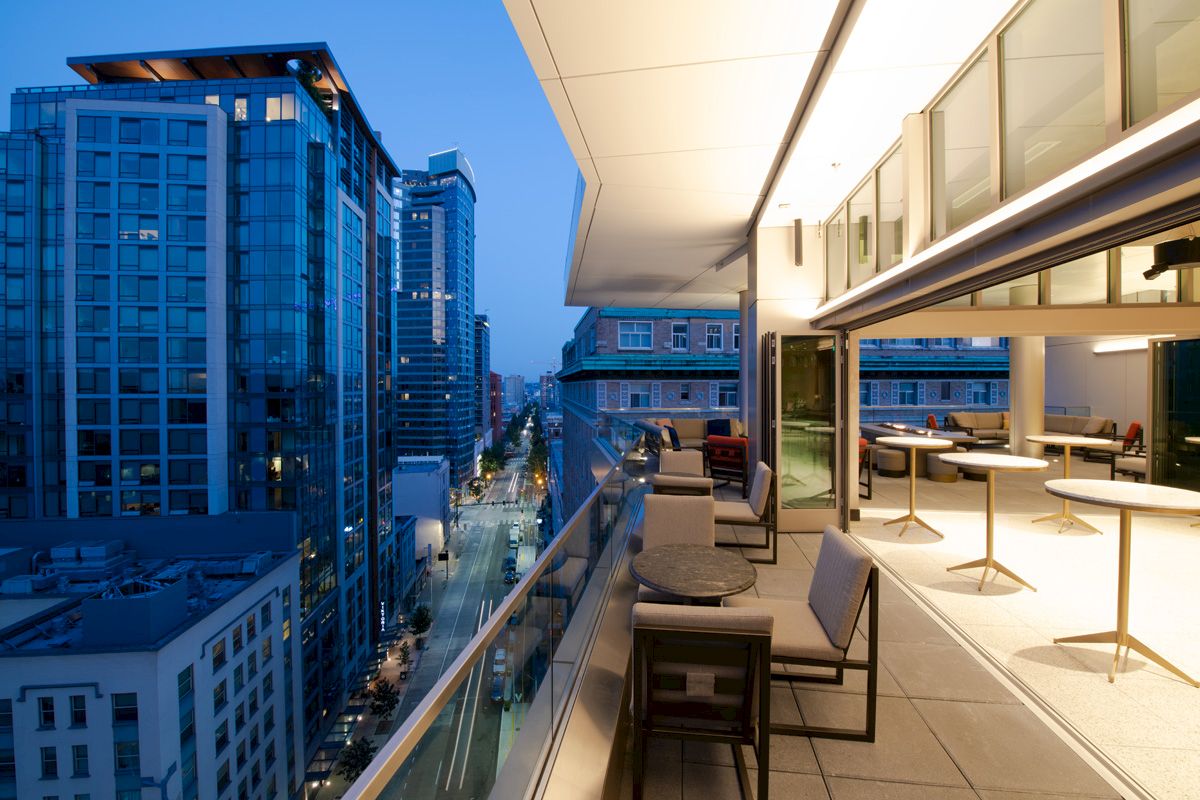 A modern rooftop terrace at dusk with seating areas, overlooking a cityscape of tall glass buildings and illuminated streets.