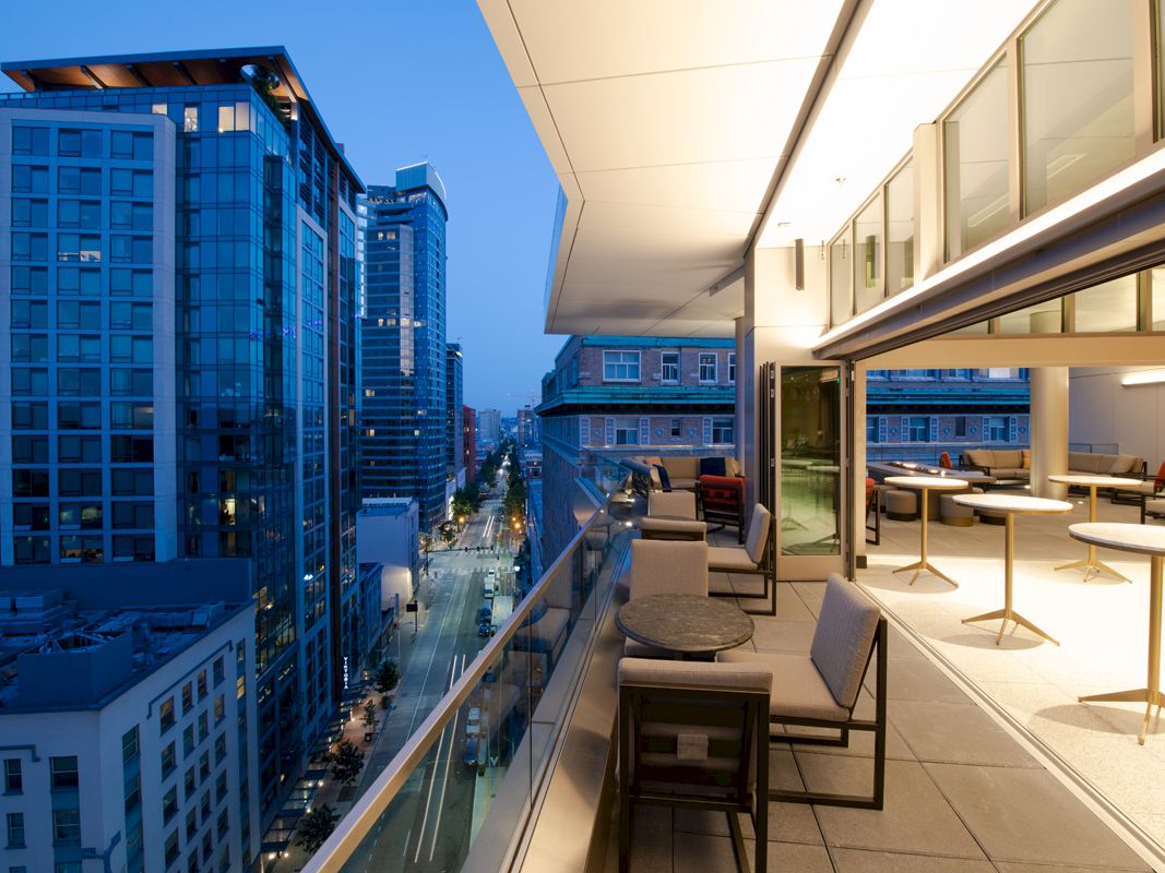 A modern rooftop terrace at dusk with seating areas, overlooking a cityscape of tall glass buildings and illuminated streets.