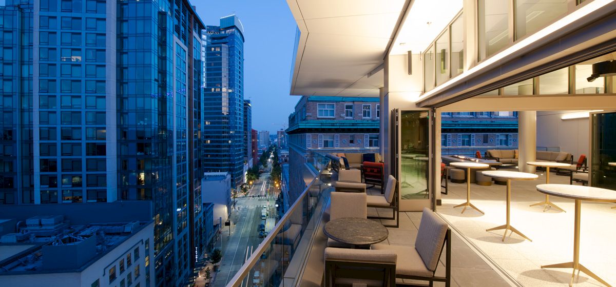 A modern rooftop terrace at dusk with seating areas, overlooking a cityscape of tall glass buildings and illuminated streets.
