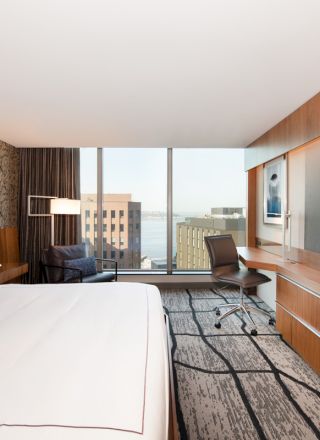 The image shows a modern hotel room with a large bed, a desk, a TV, and a window offering a city view.