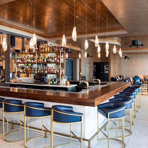 The image shows a modern bar with a wooden counter, blue stools, hanging lights, and a well-stocked back bar.