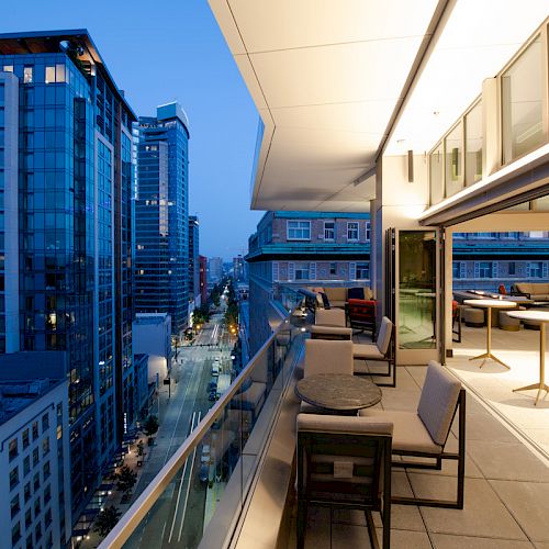 The image shows a cityscape at dusk with a modern balcony view, featuring outdoor seating and tall buildings glowing in the evening light.