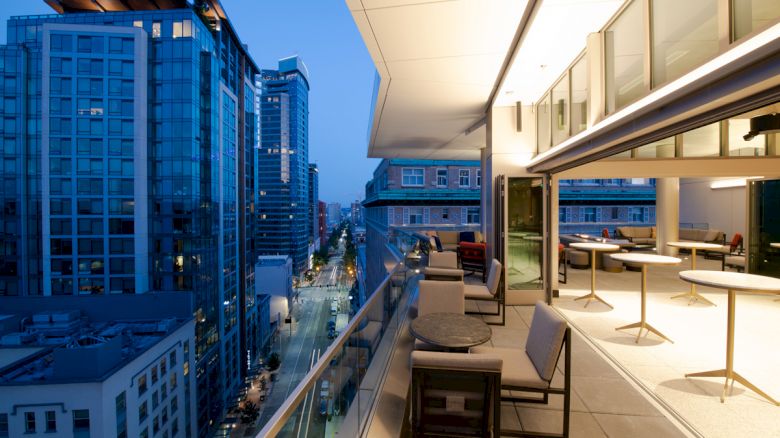 A modern rooftop terrace overlooks a cityscape at dusk, featuring outdoor seating and tables along the railing.