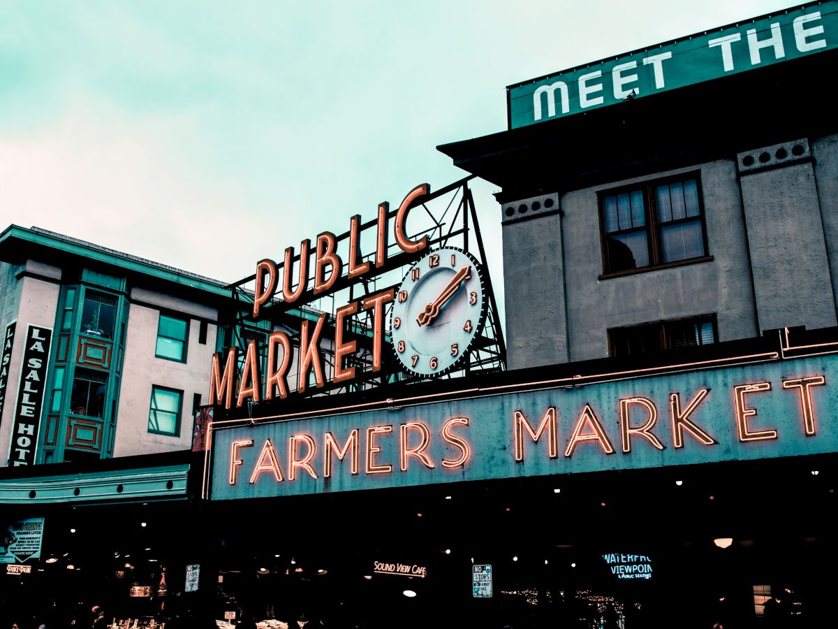 A neon sign for a "Public Market" and "Farmers Market" with a large clock is visible, set against urban buildings under a cloudy sky.