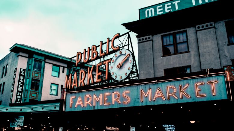 A neon sign for a "Public Market" and "Farmers Market" with a large clock is visible, set against urban buildings under a cloudy sky.