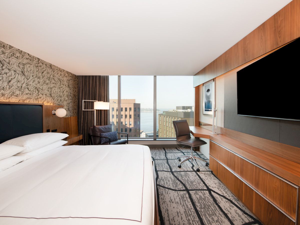 The image shows a modern hotel room with a large bed, wall-mounted TV, desk, and a city view through a window.