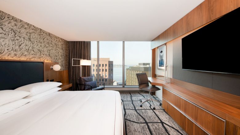 The image shows a modern hotel room with a large bed, wall-mounted TV, desk, and a city view through a window.
