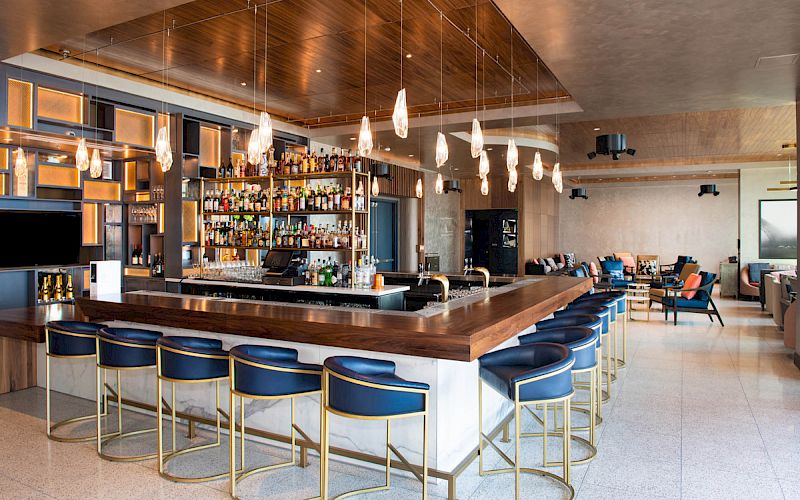 The image shows a modern bar area with a long counter, blue barstools, hanging lights, and stocked shelves in a stylish, spacious room.