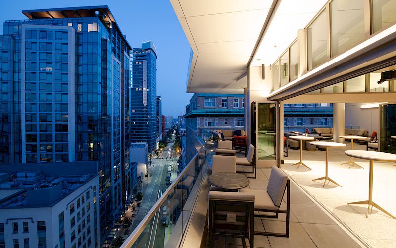 A modern rooftop patio overlooking a cityscape at dusk, with seating areas and tall buildings in the background.