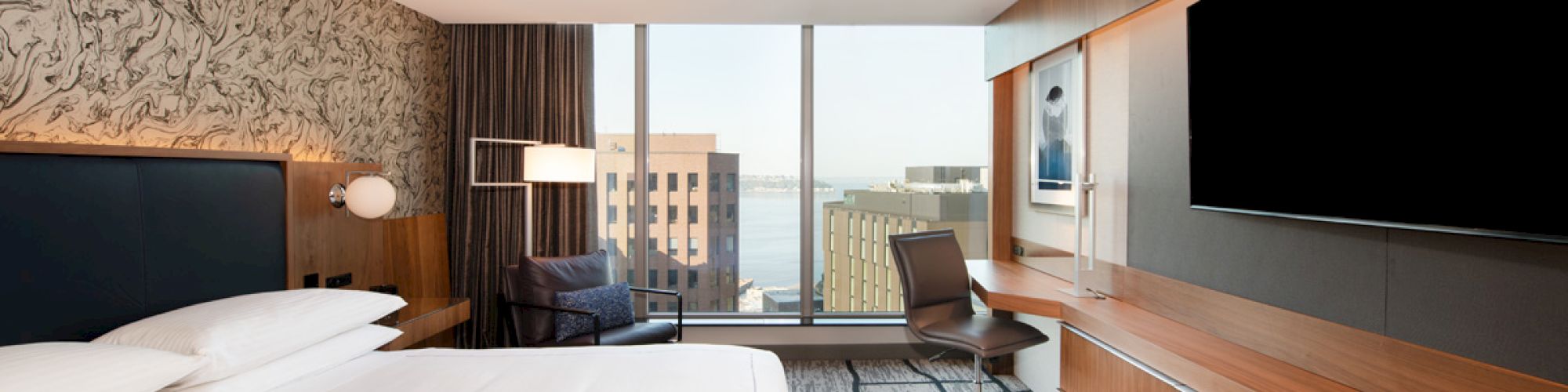A modern hotel room with a large bed, a wall-mounted TV, a desk and chair, and a window with a city view, featuring stylish decor.