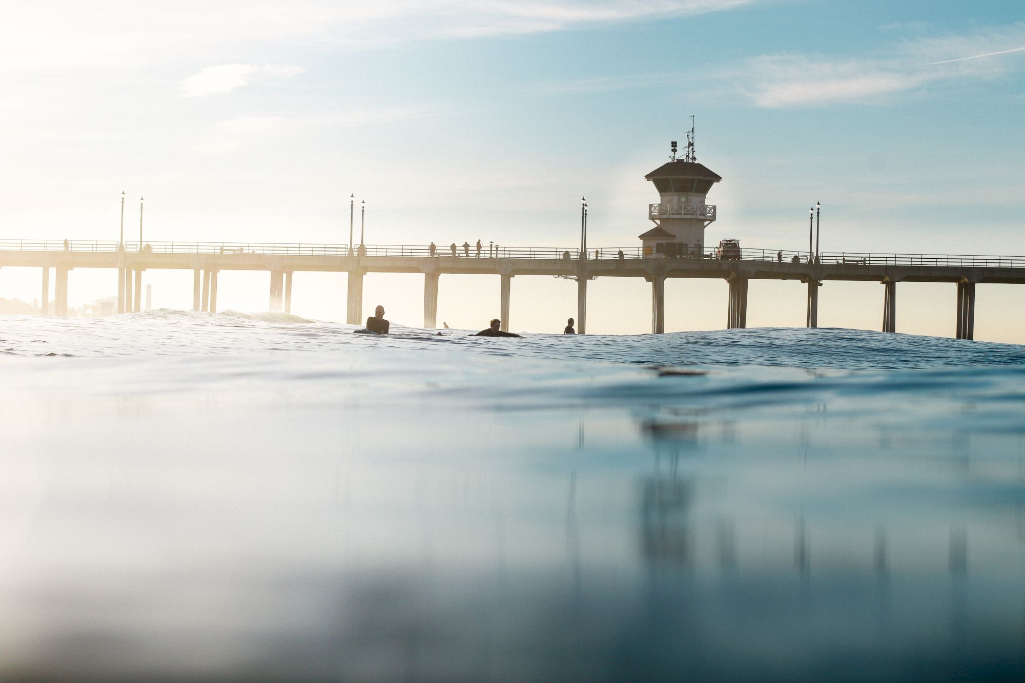 A pier stretches into the ocean with a few people on it, under a clear sky. The water in the foreground creates a serene atmosphere.